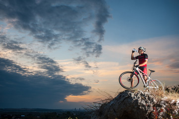Naklejka premium Cross country biker drinking water on top of a mountain with bike in the evening, sky background. side view