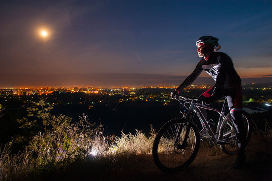 Young cyclist with mountain bike on top of the hill observing the night city view.
