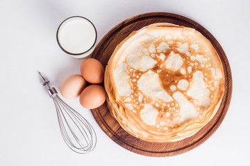 Thin pancakes on a round wooden stand with a whisk, and ingredients on a white background