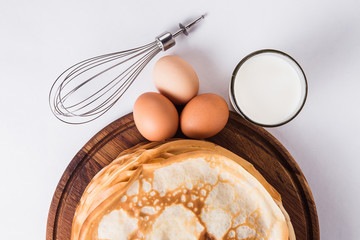 Thin pancakes on a round wooden stand with a whisk, and ingredients on a white background