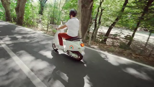 Side view of young Caucasian male riding his scooter on country road through forest