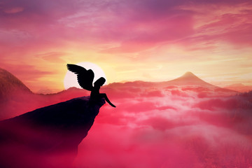Silhouette of a lonely fallen angel with long wings standing on a cliff against a paradise sunset. Dusk sky over the clouds in the mountains. Heaven landscape scene screen saver