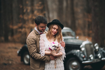 Romantic fairytale wedding couple kissing and embracing in pine forest near retro car.