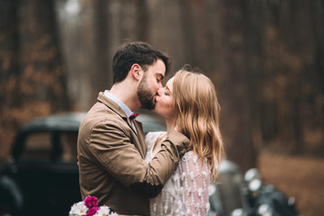 Romantic fairytale wedding couple kissing and embracing in pine forest near retro car.