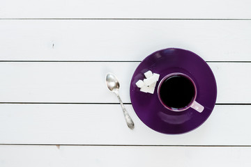 cup of tea or coffee on violet plate, silver tea spoon, lump sugar on white colored wooden table,  top view