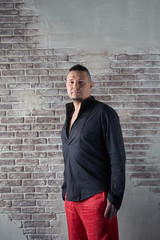 Portrait of a young man, chubby, dressed in red slacks and black shirt, hands in his pockets, fashionable hairstyle with shaved temples and slicked back hair at the top of the head, Brick wall, beard