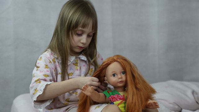 Girl playing with doll sitting on the bed, she braids her hair
