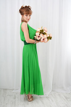  Красивая сексуальная привлекательная девушка в зеленом платье и  букетом цветов в руке позирует в белой комнате (фотостудии)

