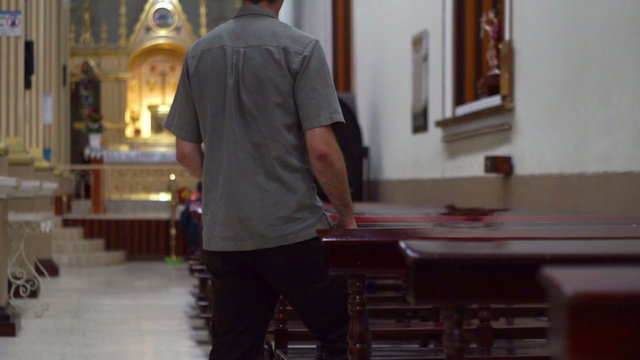 Man walks into the scene of many church benches and a distant altar and kneels on a church pew to worship and pray to his god.