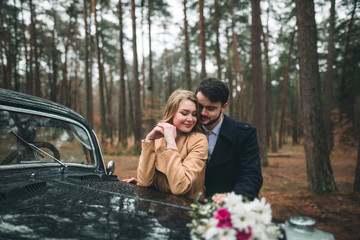 Romantic fairytale wedding couple kissing and embracing in pine forest near retro car.