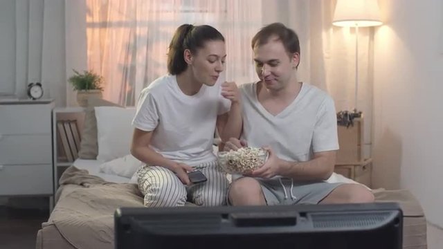 Man bringing bowl of popcorn, sitting on the bed next to his girlfriend and they start eating and watching TV