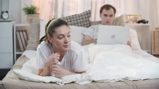 Woman holding pregnancy test and going to tell her husband that he will be a daddy while he lying on bed and using laptop