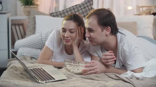 Boyfriend and girlfriend lying on bed and eating popcorn while watching interesting movie together
