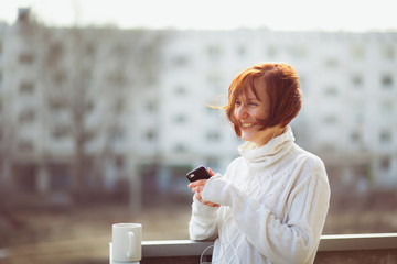 Young beautiful woman drinking tea on the balcony in a white sweater on a background of residential buildings. Still from the film.
