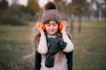 Девочка с наушниками из маленьких оранжевых тыковок слушает музыку осенью. - obrazy, fototapety, plakaty