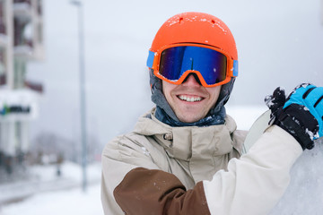 Man snowboarder wearing orange helmet, grey jacket, black and blue gloves standing with snowboard
