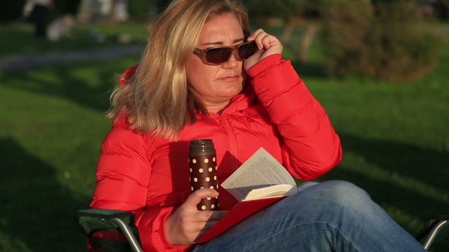 Middle age, blonde  woman reading a book  Sitting on lounge chair and dringing coffee at thermos. Resting and relaxing at the park
