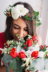 beautiful young girl in a red dress at a window with a wreath on his head and a bouquet of flowers, enjoying the smell of flowers