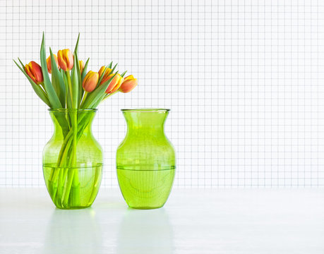 Two identical green glass vases with tulips against checkered background.