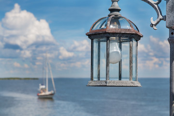 Lamp post lantern with light bulb by the ocean with sailboat horizon white fluffy clouds