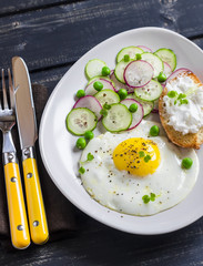 Fried egg, salad with cucumbers, radishes and green peas, toast with feta cheese on a light ceramic plate on dark wooden background. Healthy breakfast