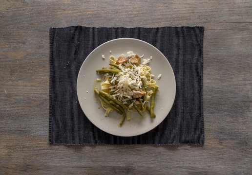 White Plate With Tagliatelle Pasta, Green Haricot Beans, Organic Chicken Meat With Grinded Cumminseed, Cream, Vermouth And Thyme And Grated Parmesan Cheese On A Black Placemat On A Wooden Table