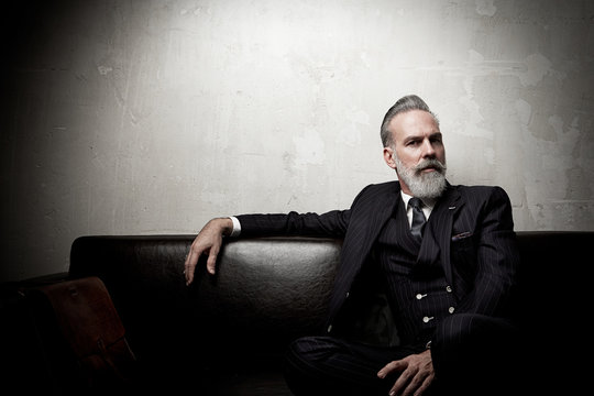 Portrait Of Adult Businessman Wearing Trendy Suit And Sitting Modern Studio On Leather Sofa Against The Empty Concrete Wall. Horizontal, Space For Ad