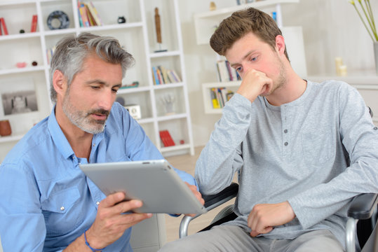 Man showing screen of tablet to young man in wheelchair