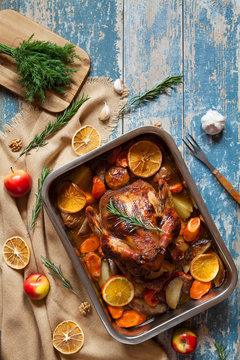 Roasted whole chicken with vegetables and herbs, rosemary and garlic in baking dish on vintage wooden table background. Top view, rustic style