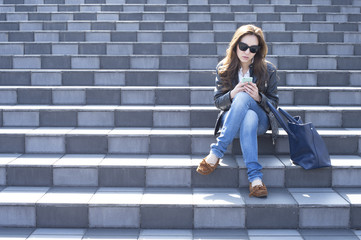 Young woman with sunglasses is looking at the smartphone sitting on the stairs