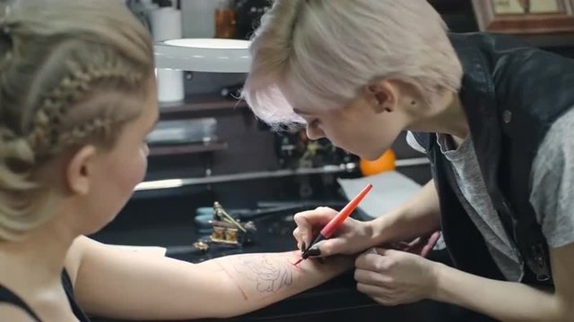 Young female tattooer using felt tip pen to draw a floral picture on arm of client 