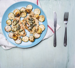 delicious, juicy chicken breast with herbs, zucchini, on a blue plate on a white napkin with knife and fork on wooden rustic background top view close up
