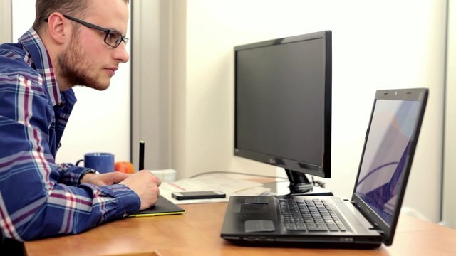 Young, handsome men sitting beind the deska and using computer and smartphone, he is little surprised and mad because of what he sees.
