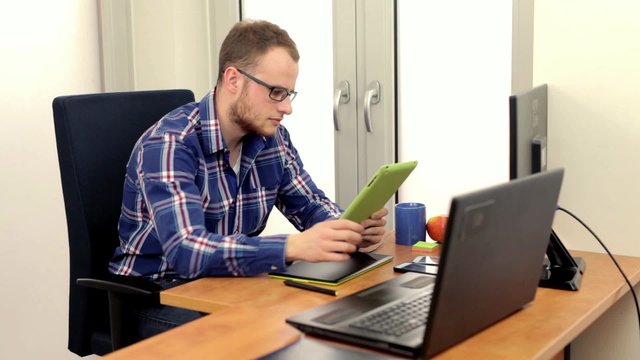 Young handsome men, siting behing his desk and alternately using tablet and smartphone, he is smiling. Positive emotions