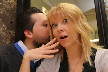 man whispers in ear to woman telling  a secret -  pretty girl looks surprised by his words