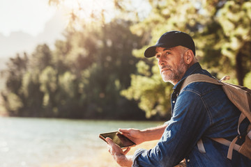 Senior man at a lake with backpack and digital tablet - Powered by Adobe