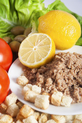 Tuna chunks served with lemon on a white plate next to the fresh vegetables, olives and croutons.