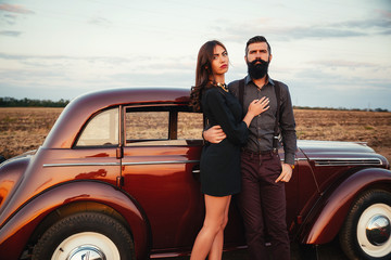 brutal bearded man with a mustache in a shirt, pants with suspenders with a girl with dark hair and big lips with bright red lipstick in a short dress and heels near retro car at sunset