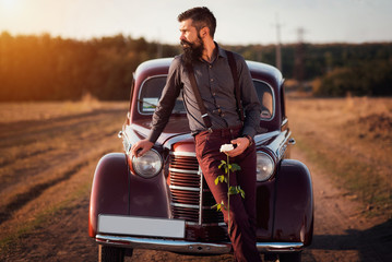 brutal bearded man with a mustache in a shirt, pants with suspenders stands leaning on the hood of...