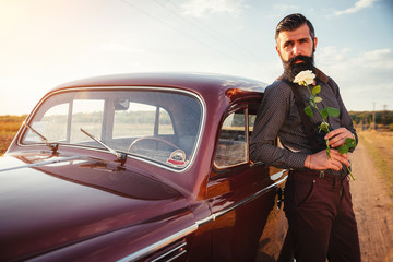 brutal bearded man with a mustache in a shirt, pants with suspenders stands leaning on the hood of a retro car holding a white rose and looking at the sunset