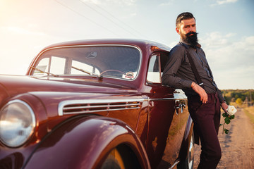 brutal bearded man with a mustache in a shirt, pants with suspenders stands leaning on the hood of a retro car holding a white rose and looking at the sunset