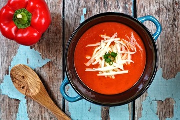 Red pepper soup topped with shredded cheese and green onions, overhead scene on rustic blue wood background