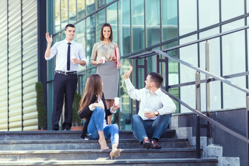Young businesswoman and businessman sitting on the steps of the office building and drinking coffee while their two colleagues are approaching and greeting them.
