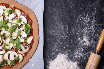 Raw pizza with vegetables on baking paper with flour and rolling pin. Wooden background, top view, copy space.