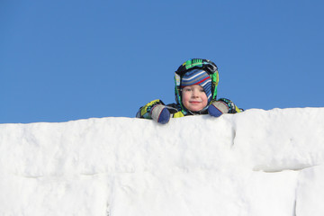 Fototapeta na wymiar The little boy in a color jacket with a hood at wall top from snow blocks 