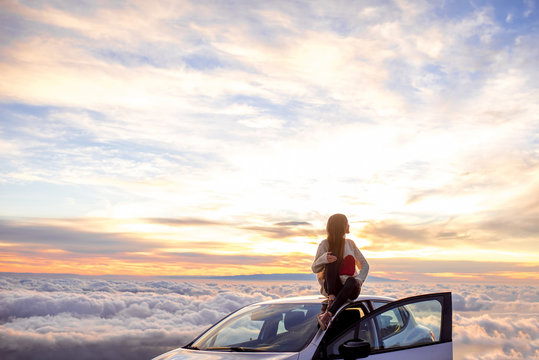 Young woman in sweater with heart shape enjoying beautiful cloudscape sitting on the car roof above the clouds on the sunrise. Wide angle image with a lot of space