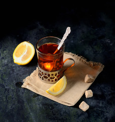 Vintage glass-holder on knitted napkin with cup of tea with sliced lemon and sugar cubes over old metal table.