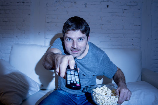 man at home lying on couch at living room watching tv eating popcorn bowl using remote control