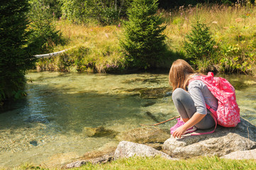 Cute little girl of 7-8 years old hiking in swiss Alps, resting by the river, wearing sport clothes, trainers and backpack, back view
