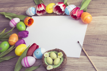 Photograph of a couple of colorful easter eggs with a bunch of tulips alligned around a sheet of white paper with copy-space on wooden surface